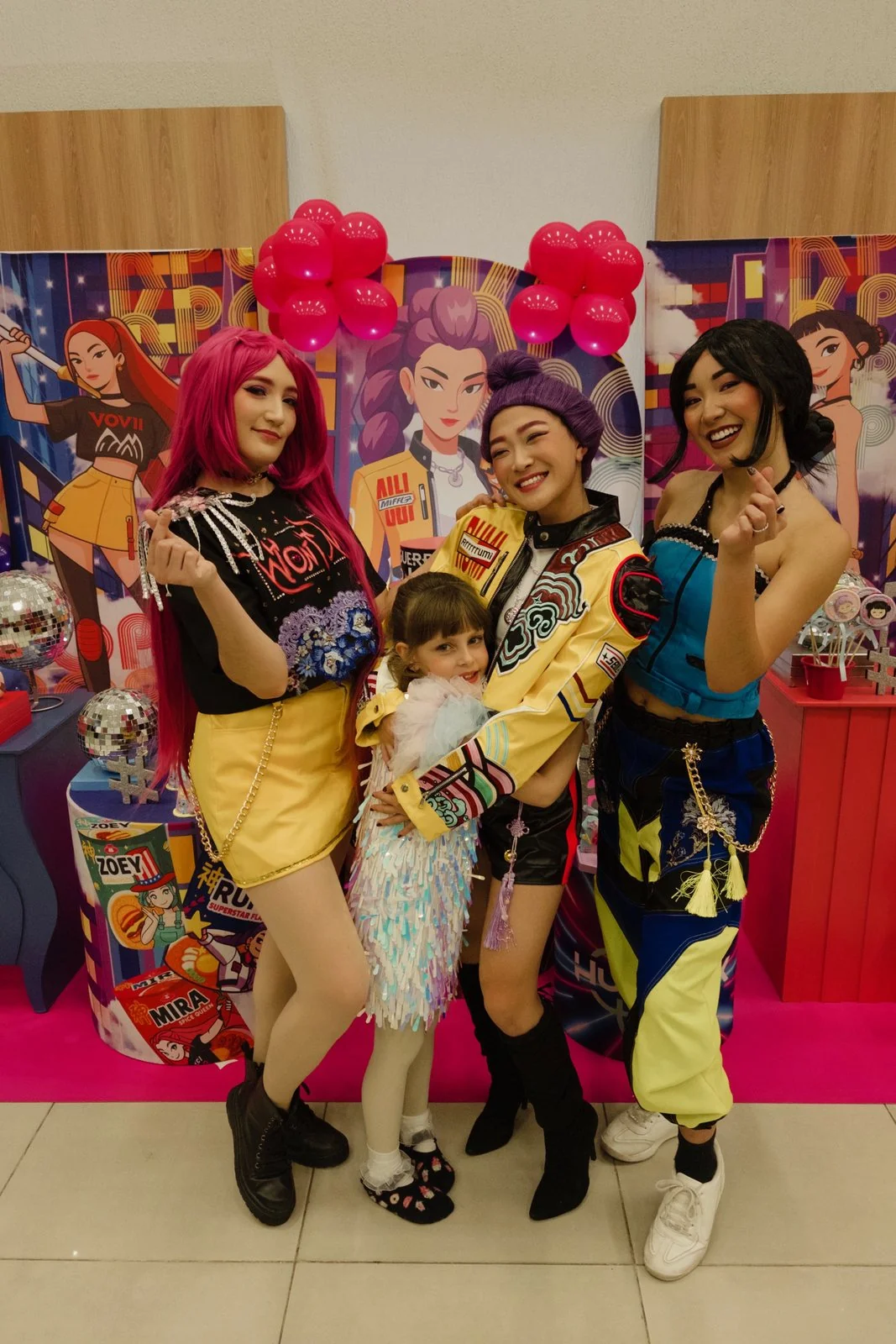 As três do K-Pop abraçadas com uma criança, diante do painel colorido da festa
